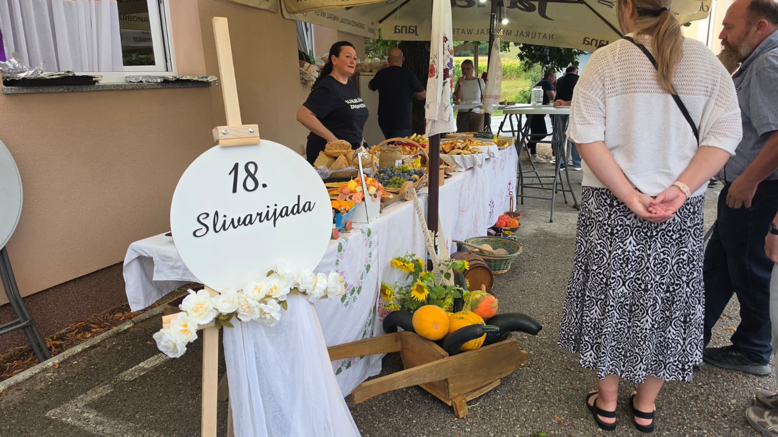 Ulaz na 18. Slivarijadu u Dropkovcu — ploča s natpisom 18. Slivarijada ispred stola s domaćim delicijama i gostima pod suncobranima