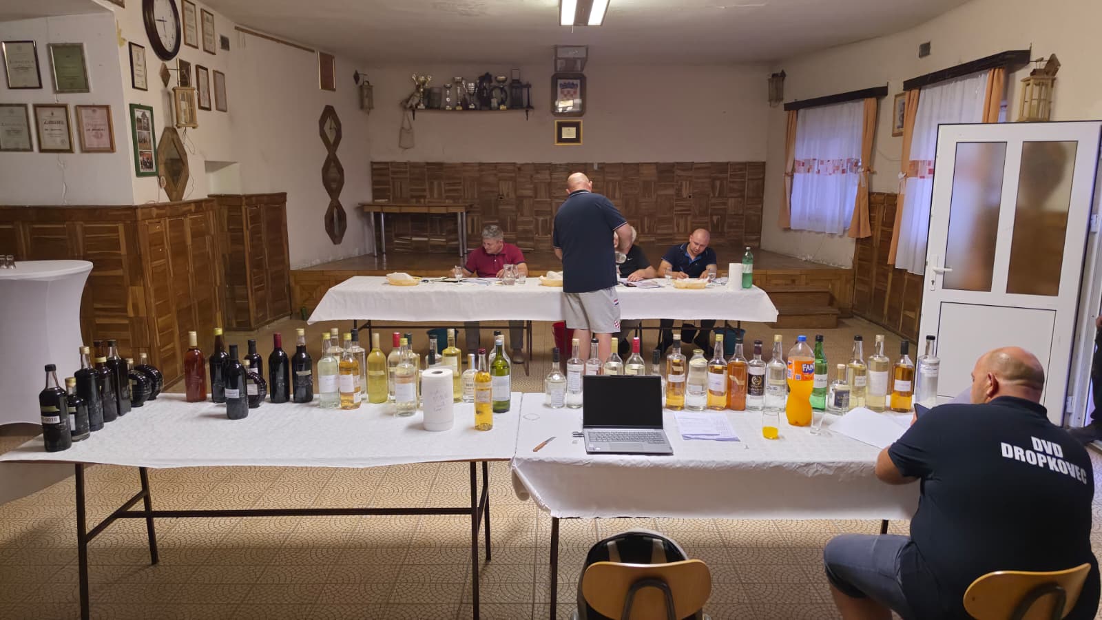 Ocjenjivanje rakija za 18. Slivarijadu u dvorani Vatrogasnog doma DVD-a Dropkovec — stolovi s numeriranim bocama uzoraka i stručno povjerenstvo u pozadini